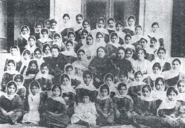 Ein Schwarz-Weiß-Foto einer Gruppe von Kindern, die vor einem Gebäude mit einer Wand und Fenstern posieren, mit dem Text "Aserbaidschanische Mädchenschule" unten.