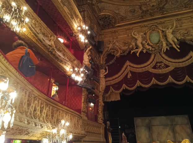 Das Innere eines Theaters mit Kronleuchtern an der Decke, Skulpturen an den Wänden und Menschen auf der Bühne, mit verstreuten Gegenständen, die eine lebendige Atmosphäre schaffen.