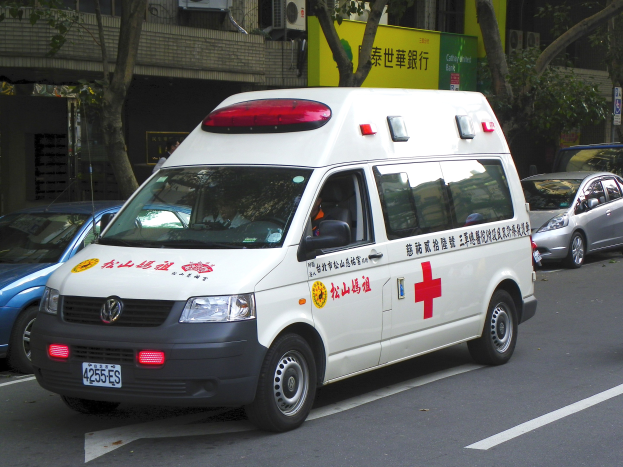 Ambulanz am Straßenrand mit anderen Fahrzeugen, Bäumen, Gebäuden, Schildern und Klimageräten im Hintergrund.