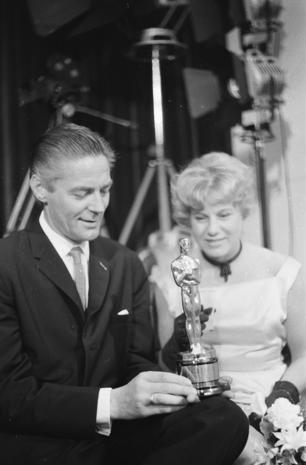 Ein Mann in einem schwarzen Blazer, weißem Hemd und Krawatte hält eine Oscar-Statue in der Hand, während er neben einer Frau in Abendgarderobe sitzt, die einen Blumenstrauß hält, beide in Schwarz-Wei├č.