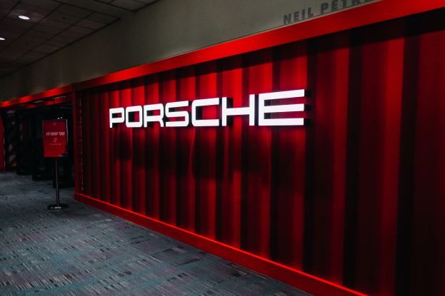 Eine rote Wand mit dem Wort "Porsche" in Deckenbeleuchtung, mit Ständern und einer Tafel auf der linken Seite, die auf eine Porsche-Ausstellung hindeutet.