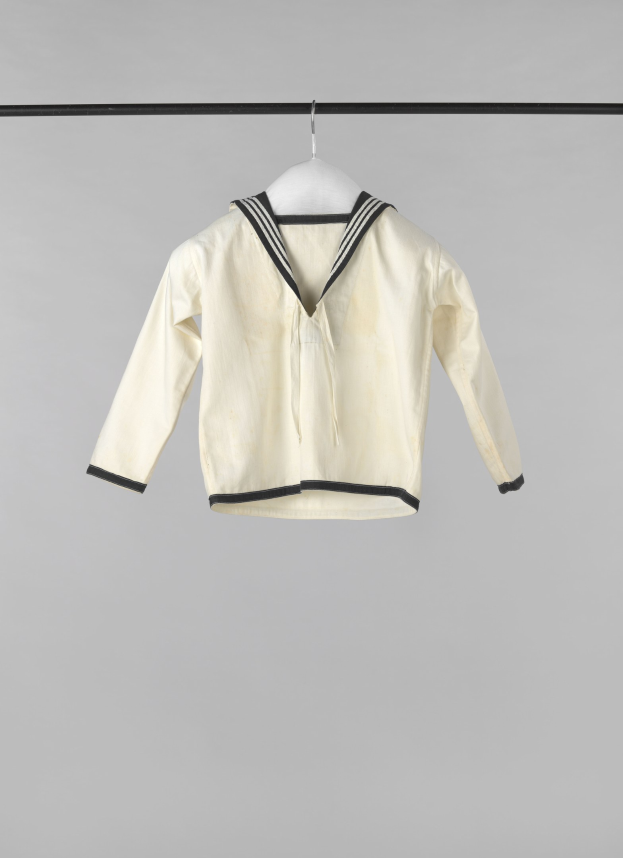 Eine weiße Matrosenjacke mit Kragen, langen Ärmeln und Knopfleiste, die an einer Wäscheleine gegen einen weißen Hintergrund hängt, aus leichtem, geripptem Stoff.