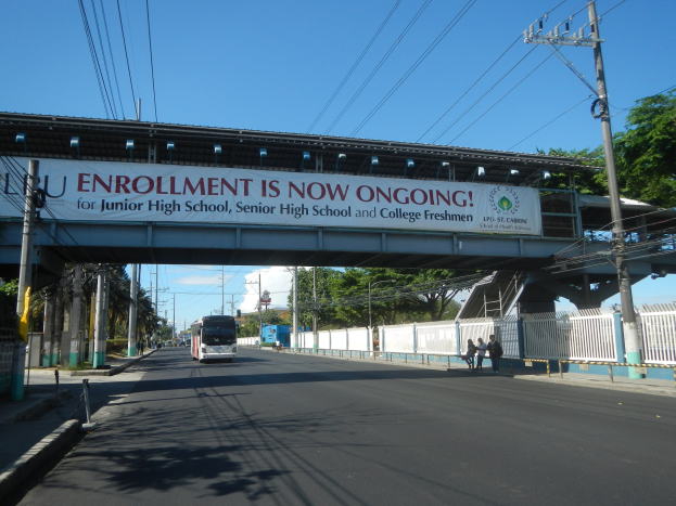 Ein Bus fährt unter einer Brücke mit einem "Jetzt Anmelden"-Schild, während Fußgänger darunter gehen und Strommasten die Straße säumen, mit Bäumen und einem blauen Himmel im Hintergrund.