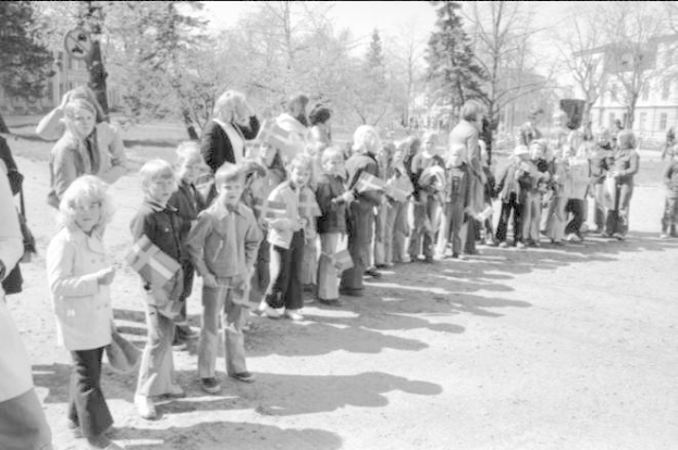 Ein Schwarz-Weiß-Bild einer Gruppe von Menschen, die in einer Reihe auf einer Schotterstraße stehen, Fahnen halten, mit Bäumen, Gebäuden und einem klaren Himmel im Hintergrund, die an einer Protestdemo auf dem Schulgelände teilnehmen.