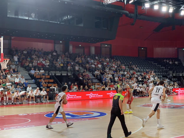 Eine Gruppe von Menschen spielt Basketball in einer Turnhalle, mit einem Basketballkorb links und Zuschauern im Hintergrund.