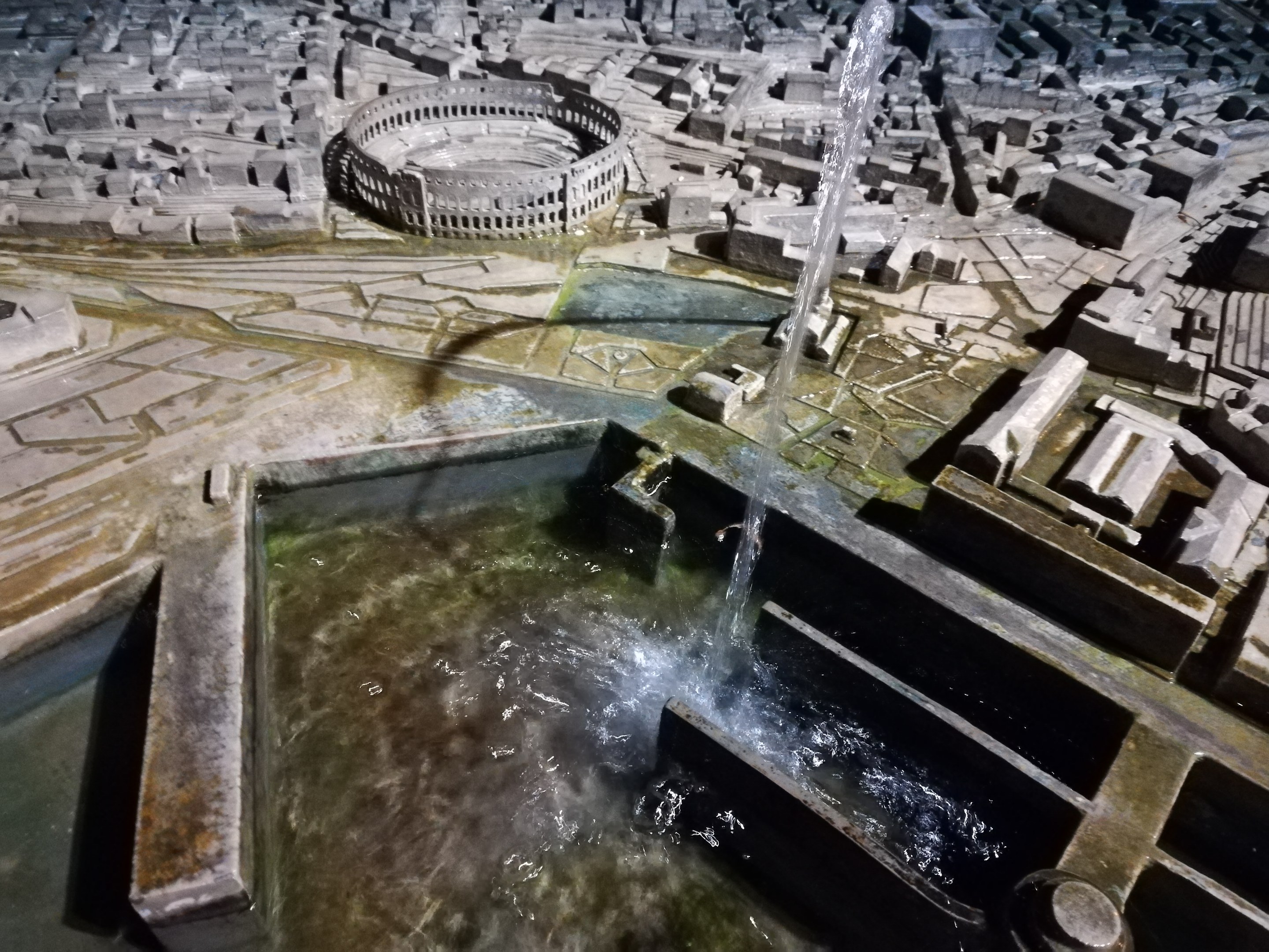 Ein Miniaturstadtmodell mit einem zentralen Brunnen mit Wasserfall, umgeben von Gebäuden und einem Gewässer.