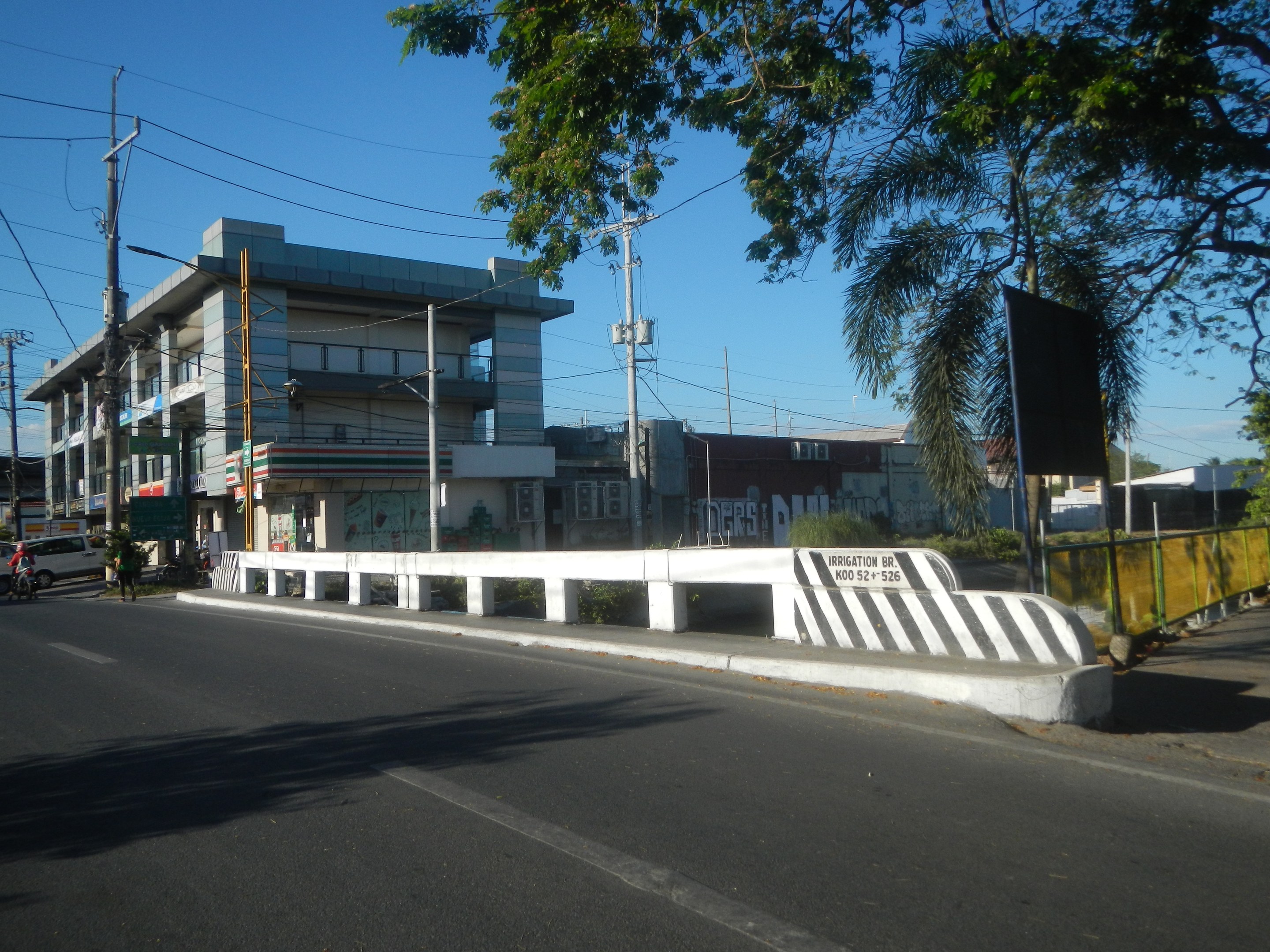 Stadtstraße mit Gebäuden, Strommasten, Kabeln, Fahrzeugen, Absperrpollern, Schildern, Bäumen, Pflanzen und einem klaren blauen Himmel.