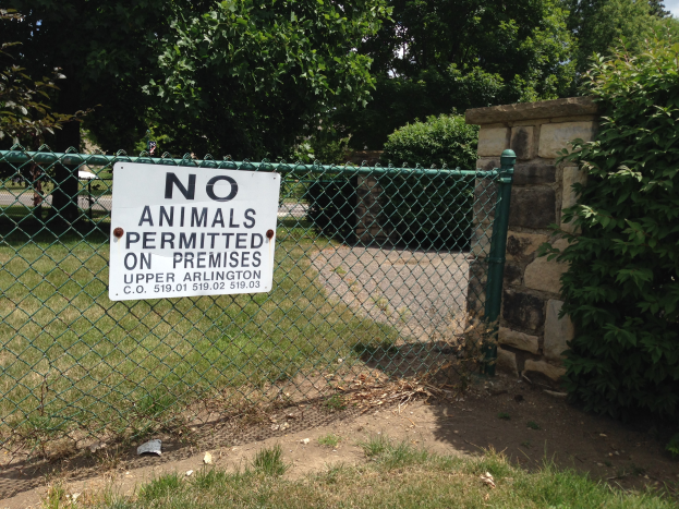 Ein Schild an einer Kettenzaun mit der Aufschrift "Keine Tiere erlaubt" umgeben von saftigem Grün, Pflanzen und Bäumen, mit einer Wand im Hintergrund und einem klaren blauen Himmel darüber.