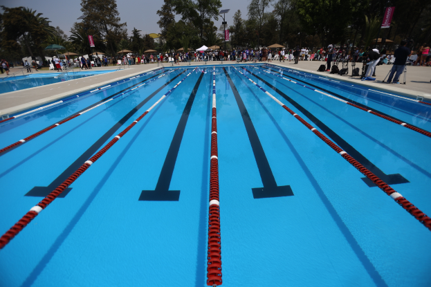 Ein großes Schwimmbad mit Bahnenteilung, umgeben von Menschen auf dem Boden, einige halten Taschen und andere Gegenstände, mit Zelten, Textbannern, Pfählen, einer Straßenlaterne, einem Schild und einem bewölkten Himmel im Hintergrund.
