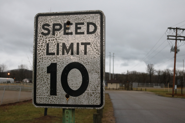 Tempolimitschild am Straßenrand mit Gras, Strommasten, einem Zaun, Häusern, Bäumen und einem bewölkten Himmel im Hintergrund.