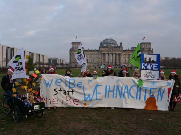 Gruppe von Menschen in Mützen, die ein Banner vor dem Reichstag halten, mit einer Person in einem Kinderwagen, Rasen, Bäumen, Gebäuden und bewölktem Himmel.