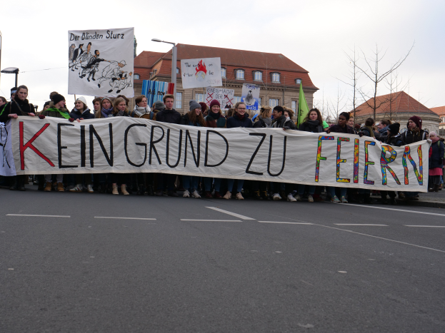 Eine Gruppe von Menschen steht auf einer Straße und hält ein Transparent mit der Aufschrift "Kein Grund zu Feiern" im Protest, mit Straßenlaternen, Bäumen, Gebäuden mit Fenstern und einem klaren blauen Himmel im Hintergrund.