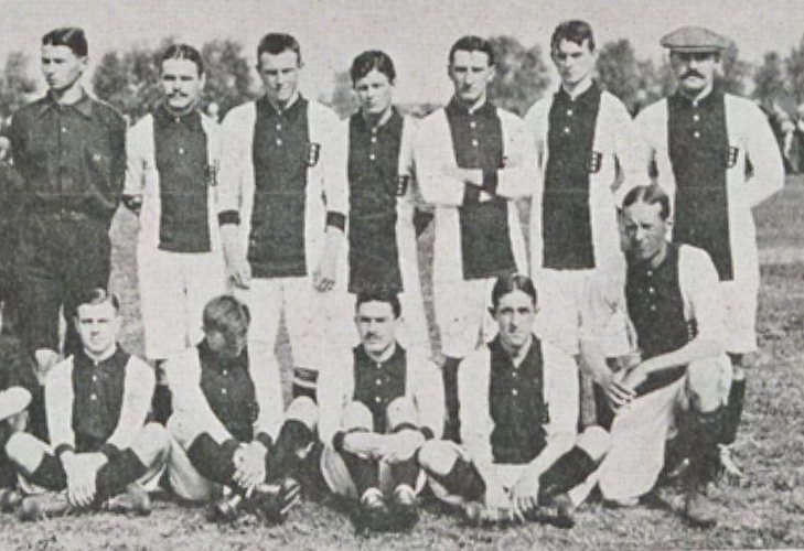 Ein Schwarz-Weiß-Foto einer Fussballmannschaft, mit einigen stehenden und anderen sitzenden Mitgliedern, sowie einigen Menschen, Bäumen und dem Himmel im Hintergrund und Text unten.