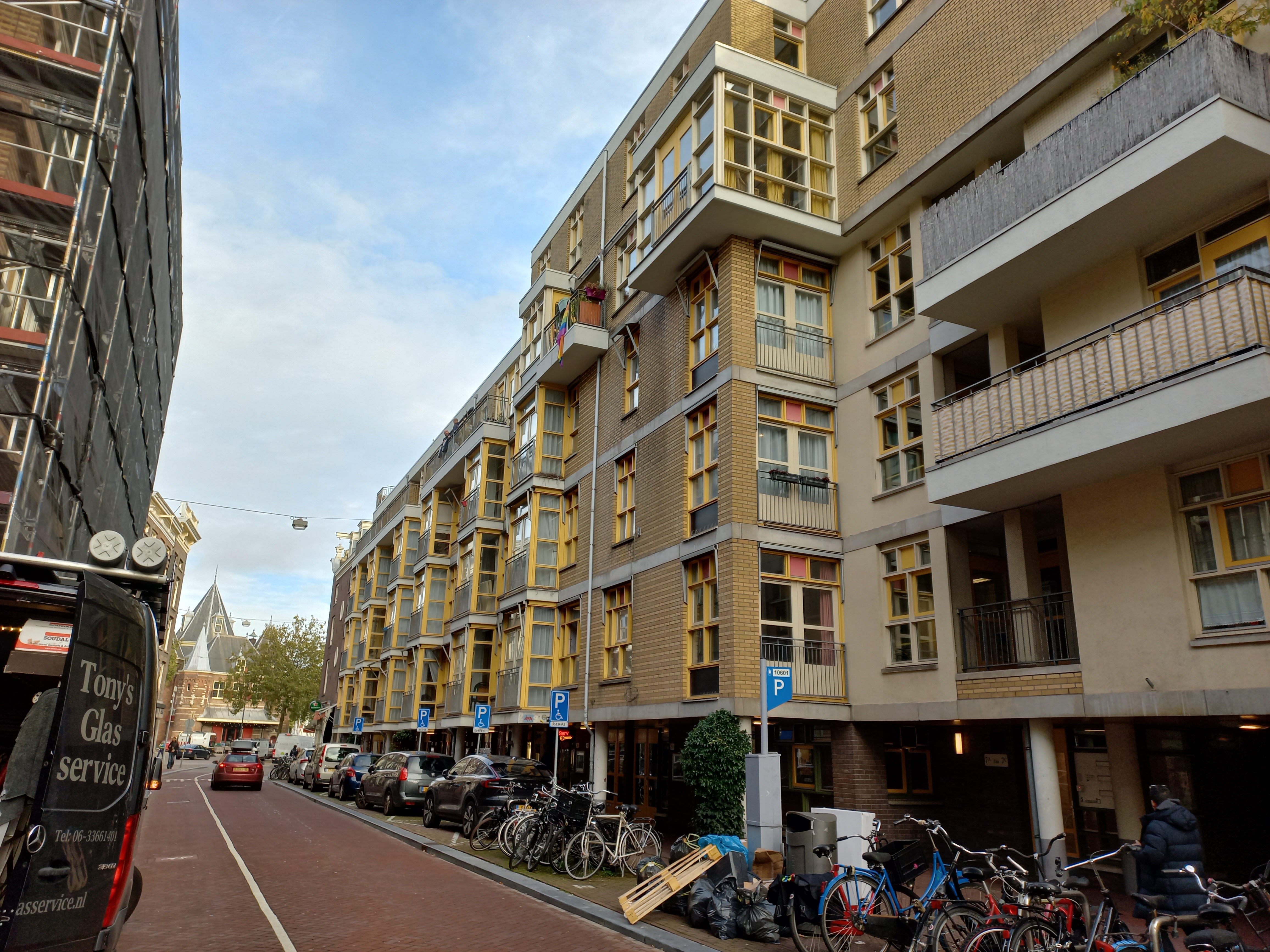 Eine Straße in Amsterdam mit parkenden Fahrrädern, fahrenden Fahrzeugen, Gebäuden mit Fenstern, Schildern an Pfählen, Bäumen und einem bewölkten Himmel.