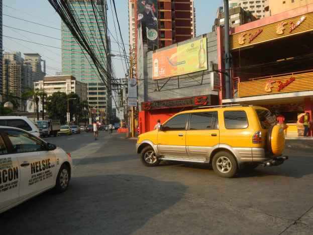 Ein gelbes Taxi fahrt eine Stra├če mit hohen Geb├Ąuden, Menschen auf dem Gehweg, Strommasten mit Kabeln und B├Ąumen entlang.