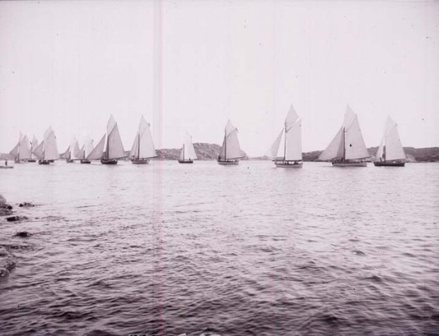 Schwarzes und weißes Foto von Segelbooten auf dem Wasser mit Hügeln im Hintergrund, klarem Himmel darüber und 'New York Yacht Club'-Text unten.