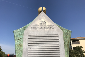 Ein hohes Denkmal mit einer Uhr darauf, mit Text beschriftet, steht in einem Park umgeben von Bäumen und Gebäuden unter einem klaren Himmel, zum Gedenken an den 24. Jahrestag des Völkermords in Armenien.