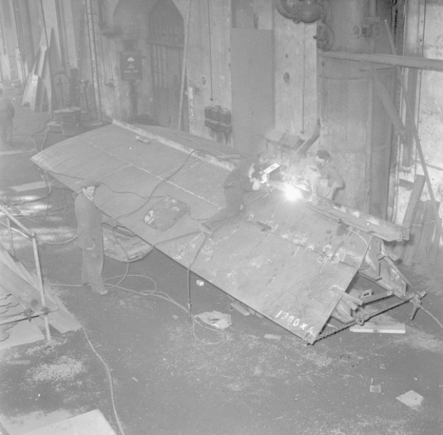 Schwarzes und weißes Bild von Männern in einer Fabrik, die an einem Metallobjekt arbeiten, einer verwendet eine Schweißmaschine.