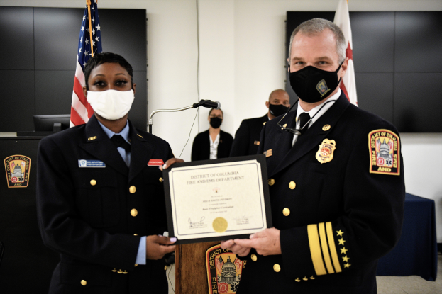 Zwei Männer in Uniform und Masken stehen nebeneinander und halten ein Zertifikat, mit einem Podium mit einem Mikrofon dahinter, Fahnen und einer Wand im Hintergrund, und Tischen mit verschiedenen Gegenständen auf beiden Seiten.