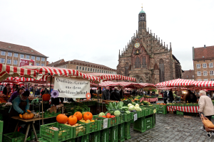 Ein lebendiger Markt in Nürnberg, Deutschland, voller verschiedener Früchte und Gemüse, Menschen mit Taschen und Zelten, mit Gebäuden und einem Kirchturm im Hintergrund unter einem klaren Himmel.