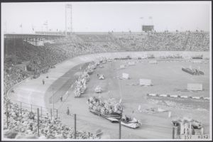 Schwarzes und weißes Foto einer Rennstrecke mit Zuschauern in einem Stadion, einige Fahrzeuge auf der Straße und Fahnen im Hintergrund.