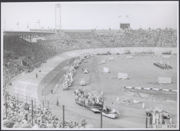 Schwarzes und weißes Foto einer Rennstrecke mit Zuschauern in einem Stadion, einige Fahrzeuge auf der Straße und Fahnen im Hintergrund.
