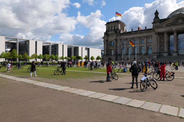 Eine Gruppe von Menschen, die vor dem Reichstaggebäude in Berlin, Deutschland, Fahrrad fährt, das mit Flaggen und Säulen geschmückt ist, mit einem bewölkten Himmel und einem grasbewachsenen Boden mit Bäumen im Hintergrund.