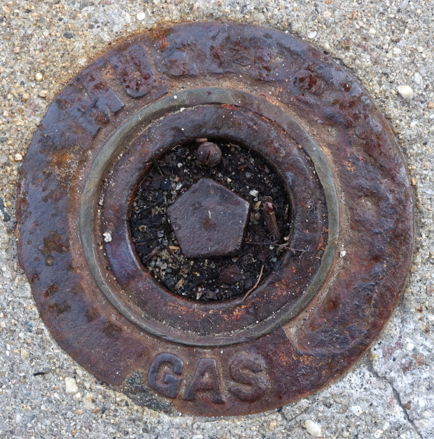 Ein verrosteter Metall-Schachtobdeckel auf dem Boden mit dem Wort "Gas" darauf.