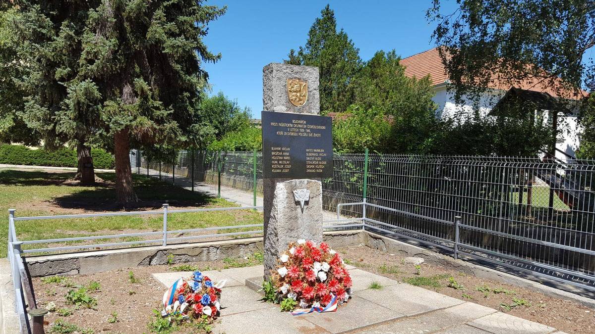 Ein Denkmal für die Opfer des Tschernobyl-Unfalls, das sich in einem Park mit Kränzen befindet, umgeben von einem Metallzaun, Gras, Pflanzen, Bäumen und einem Haus mit Dach und Fenstern, unter einem bewölkten Himmel.