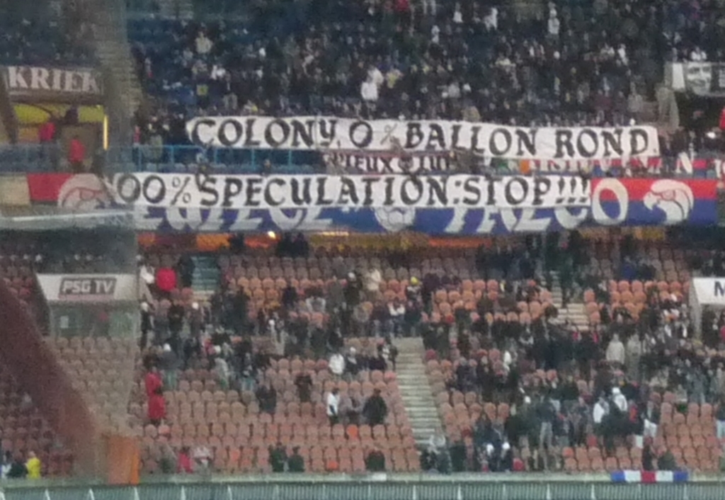 Eine große Menschenmenge in einem Stadion mit Transparenten und Stühlen, einem Zaun unten und einer Treppe links.