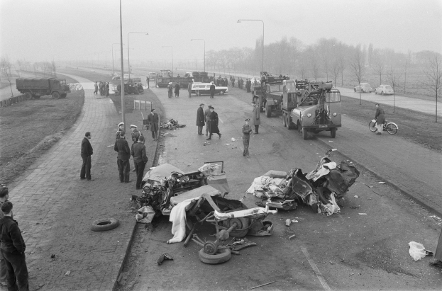 Eine Schwarz-Weiß-Szene eines Autounfalls am Straßenrand mit mehreren Fahrzeugen, darunter ein Landfahrzeug, und einer Gruppe von Menschen, die darum herumstehen, mit Laternen, Bäumen und Himmel im Hintergrund.
