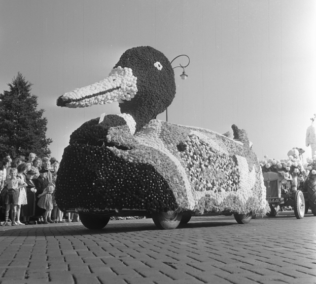 Ein Schwarz-Weiß-Foto eines Entenwagens, der mit Blumen geschmückt ist und von Menschen umgeben ist, die auf der linken Seite stehen, Bäume und einen klaren Himmel im Hintergrund.
