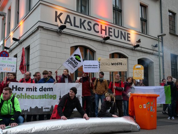 Eine Gruppe von Menschen, die vor einem Gebäude in Deutschland protestieren und Schilder und Plakate halten, mit zwei Personen im Vordergrund und einem Müllcontainer rechts.
