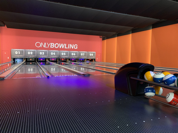 Eine Bowlinganlage mit Bowlingkugeln auf der rechten Seite, aufgestellte Kegel in der Mitte und eine Wand mit Text im Hintergrund, unter einer sichtbaren Decke.