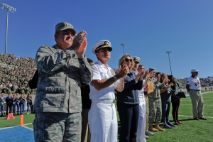Eine Gruppe von Menschen auf einem Fußballfeld klatschen, mit Zuschauern auf den Tribünen, Laternenmasten und einem klaren blauen Himmel im Hintergrund.