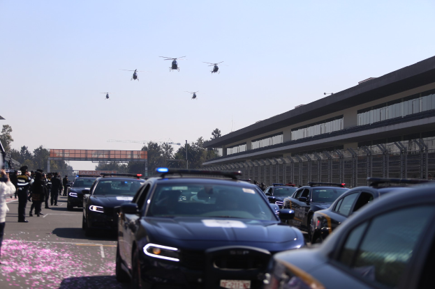 Polizeifahrzeuge vor einem Gebäude mit Menschen auf der linken Seite, Bäumen und einer Brücke im Hintergrund sowie vier Hubschraubern am klaren blauen Himmel.