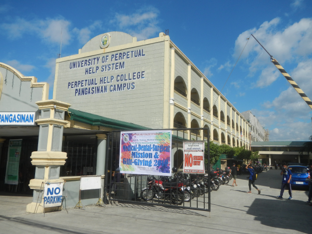Die Universit├Ąt Perpetual Help System im Pangasinan Campus, mit einer Gruppe von Menschen auf dem Boden, einem Metallzaun, einer Fahne mit Text, geparkten Fahrzeugen, einem Schild, Pfosten, einem Kran, B├Ąumen und einem bew├Âlktem Himmel.