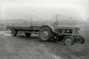 Ein Schwarz-Weiß-Foto eines Traktors, der einen Anhänger auf einem Schotterweg zieht, umgeben von Gras, Pfählen, Drähten, Bäumen, Gebäuden, Hügeln und einem klaren Himmel.