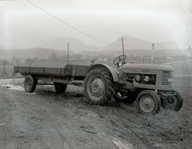 Ein Schwarz-Weiß-Foto eines Traktors, der einen Anhänger auf einem Schotterweg zieht, umgeben von Gras, Pfählen, Drähten, Bäumen, Gebäuden, Hügeln und einem klaren Himmel.