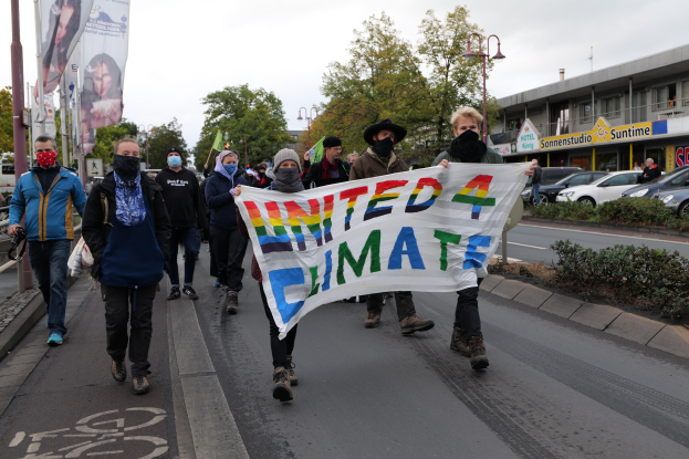 Eine Gruppe von Menschen geht eine Straße entlang, hält eine 'United 4 Climate'-Schlagzeile, trägt Masken und trägt Taschen, mit einem Gehweg, Geländer, Schlagzeilen, Bäumen, Laternenpfählen, Fahrzeugen, Gebäuden und einem klaren blauen Himmel im Hintergrund.
