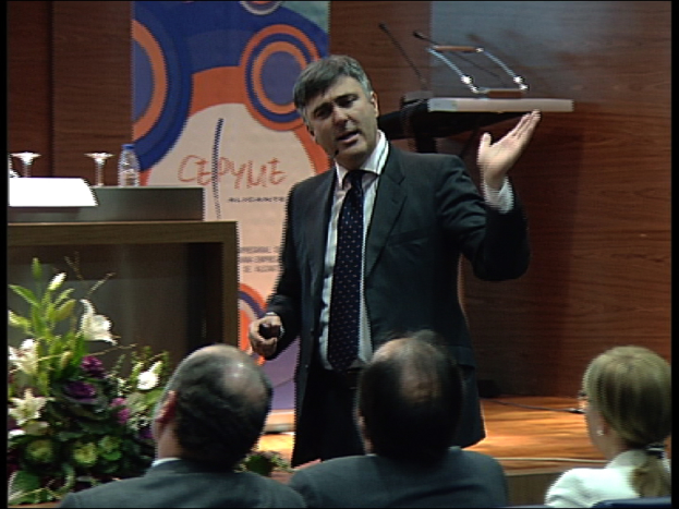 Ein Mann in einem schwarzen Blazer steht an einem Podium mit Mikrofonen, spricht in ein tragbares Mikrofon, mit einer Blumenanordnung und einem Tisch mit einem Buch und einer Wasserflasche daneben, während drei Personen auf Stühlen davor sitzen, vor einer braunen Wand und einer bunten Tafel mit Text.