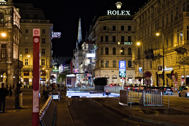 Eine nächtliche Stadtstraße mit fahrenden Autos, Fußgängern auf den Gehwegen, parkenden Fahrrädern, Straßenlaternen, hohen Gebäuden, Absperrungen, Pfählen und einem Rolex-Logo, das auf einem Gebäude im Hintergrund zu sehen ist.