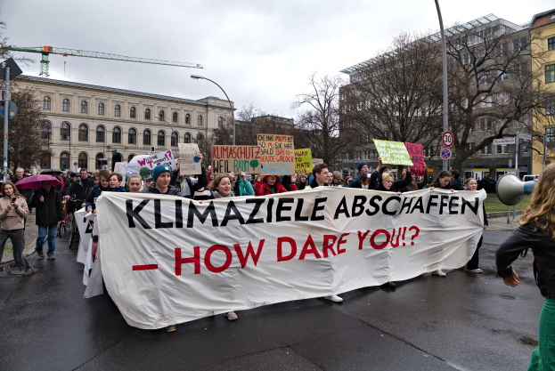 Eine Gruppe von Menschen marschiert mit einem Transparent, auf dem "Klimaziele Abschaffen - Wie wagst du es?" steht, durch die Straße. Einige halten Schirme und einer verwendet ein Megafon, vor dem Hintergrund von Laternenpfählen, Schildern, Bäumen, Gebäuden mit Fenstern, einem Kran und einem klaren blauen Himmel.
