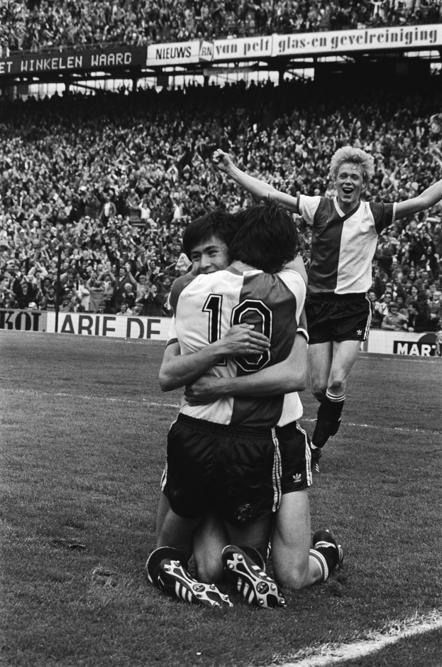 Drei Fußballspieler feiern ein Tor auf dem Boden, mit jubelnder Menge und schwenkenden Fahnen in einem schwarz-weißen Stadion.