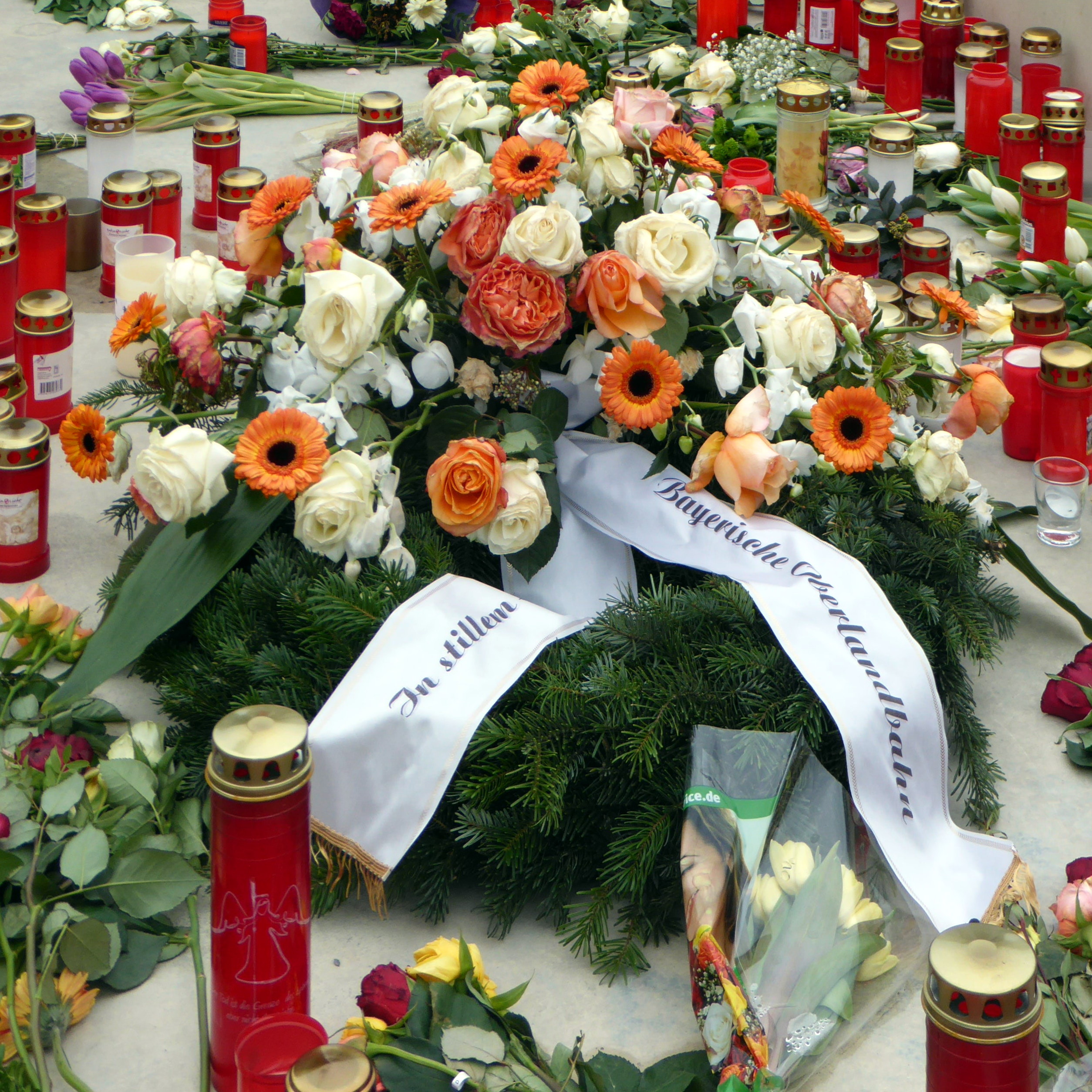 Ein Denkmal in Berlin, Deutschland, mit einem Blumenstrauß und Kerzen, die auf dem Boden vor einer Wand aufgestellt sind, umgeben von Blättern und anderen Gegenständen.