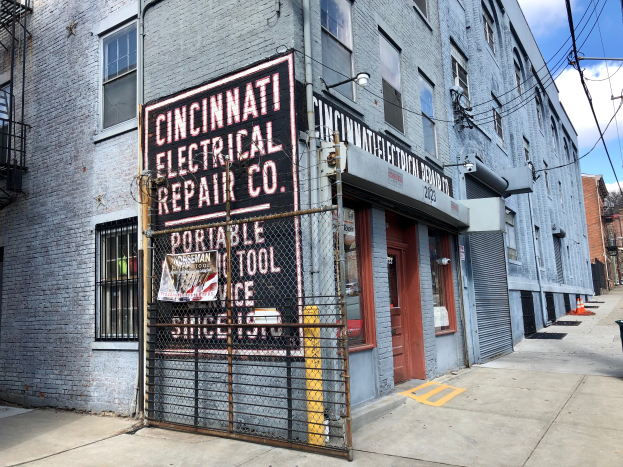 Das Cincinnati Electrical Repair Co. Gebäude mit Fenstern, Türen, einer Schautafel, einem Metallzaun, einem Müllcontainer, Polen, Drähten, Bäumen und einem bewölkten Himmel.