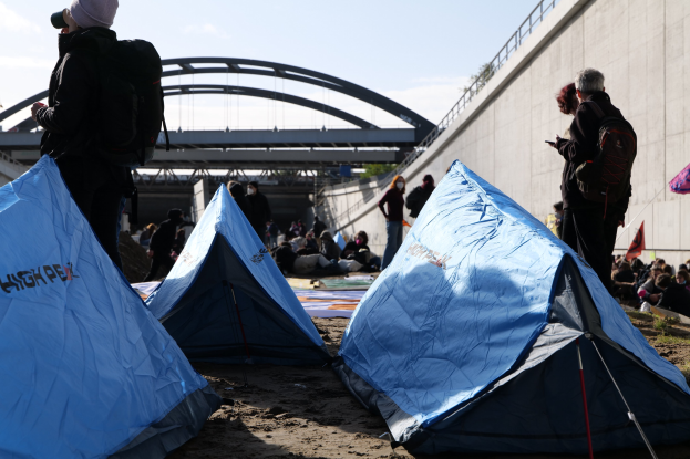 Eine Gruppe von Menschen sitzt auf einem sandigen Strand in der Nähe von Zelten, mit einer Wand und einer Brücke im Hintergrund, die an einer Klimademonstration unter einem bewölkten Himmel teilnehmen.