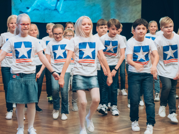 Gruppe von Kindern in weißen Hemden und blauen Shorts, die auf einem Holzboden während eines Kunstnädler-Tanzwettbewerbs tanzen, mit einem Bildschirm an der Wand im Hintergrund.