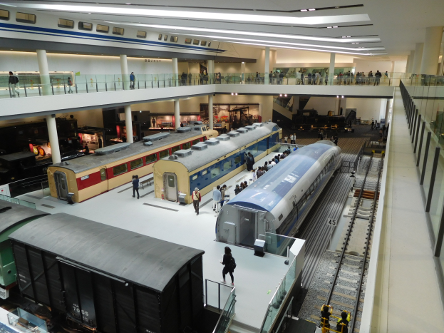 Ein geräumiger Museumsraum voller verschiedener Züge auf Display, Menschen die beobachten, Geländer, Säulen und Deckenleuchten.