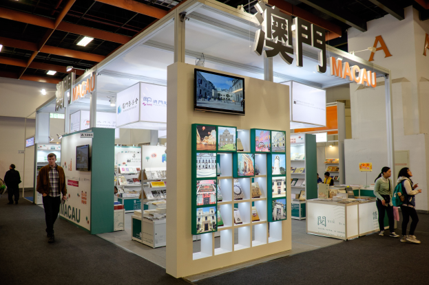 Eine Gruppe von Menschen steht vor einem Messestand auf der Macau International Book Fair, der mit Büchern, anderen Gegenständen und einem montierten Fernseher gefüllt ist, während Namensschilder und Lichter den Bereich beleuchten.